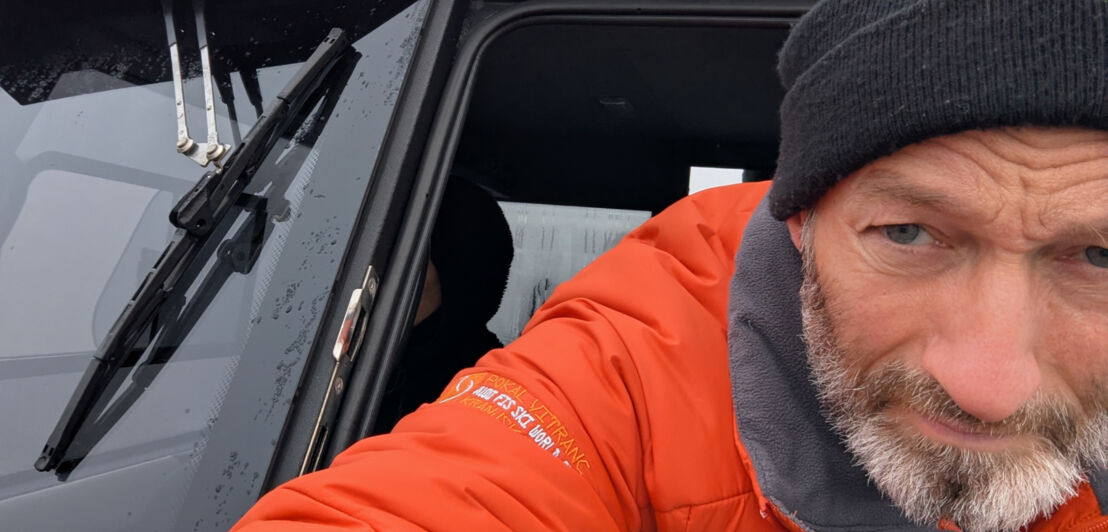 Person mit wetterfester Kleidung im Steuerhaus eines Motorboots mit regennassen Scheiben.