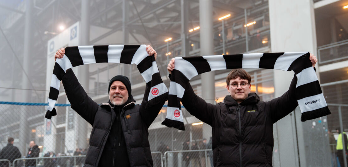 Zwei Gewinner vor dem Volksparkstadion halten Fanschals der Eintracht über ihren Köpfen hoch.