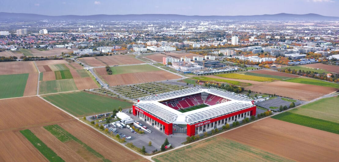 Luftaufnahme von einem rechteckigen Fußballstadion in den Farben Weiß und Rot auf einem Feld am Rande einer Großstadt.