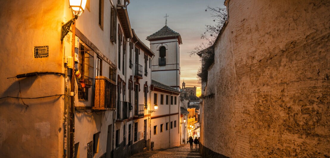 Altstadtgasse zwischen weißen Häusern und einer hohen Mauer in der Abenddämmerung.