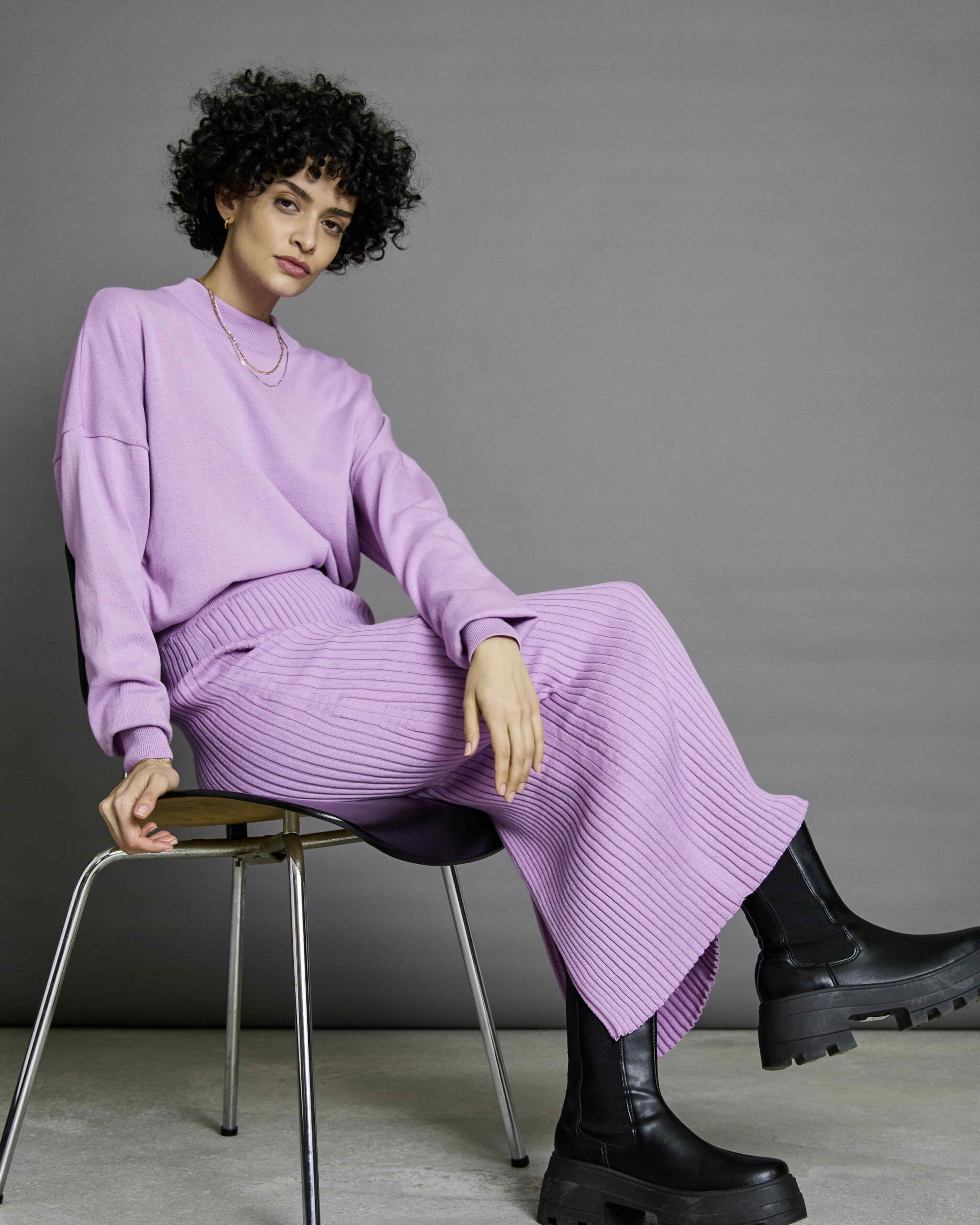 Ein Model mit schwarzen Haaren sitzt auf eine Stuhl und präsentiert fliederfarbene Mode