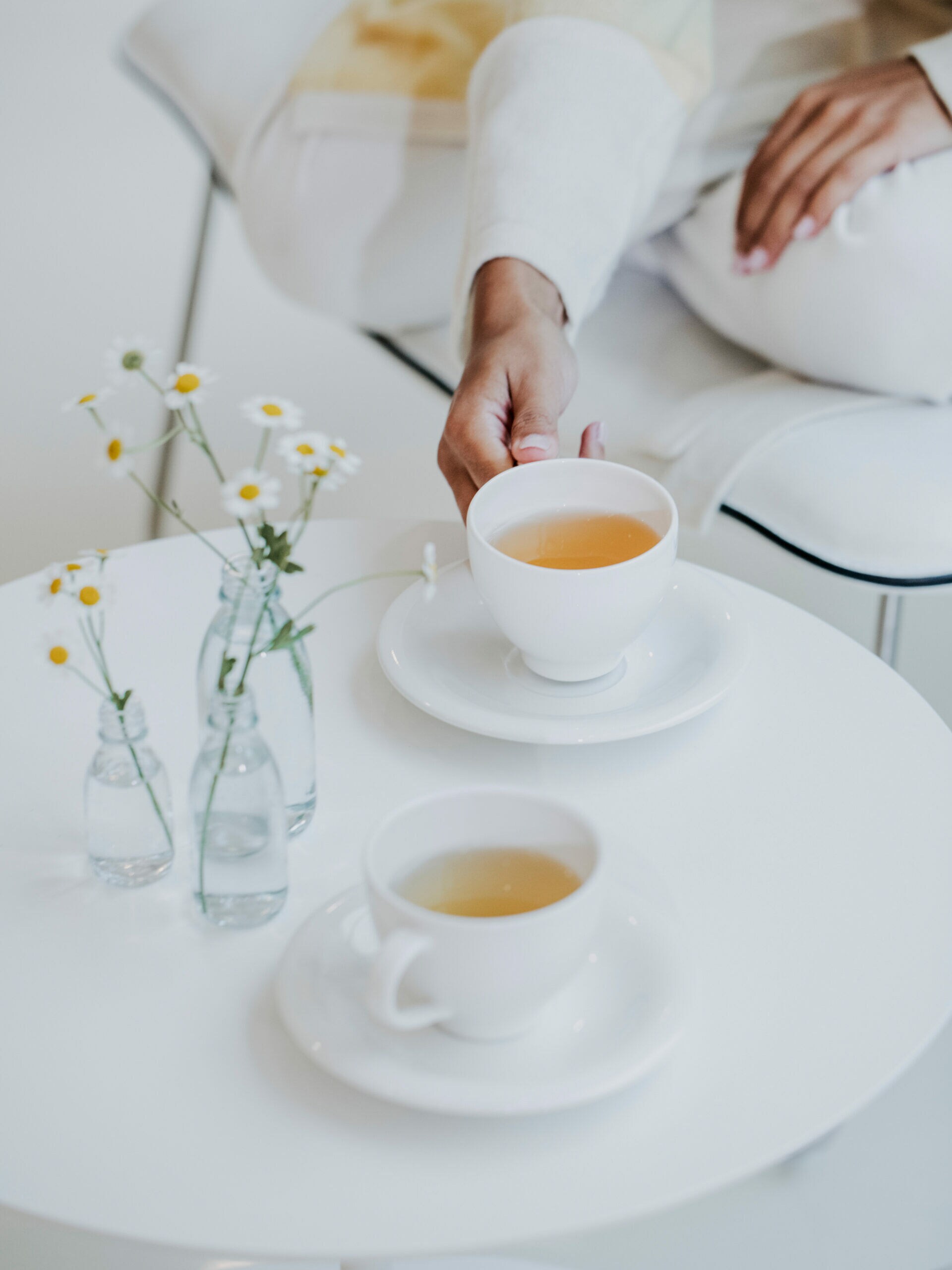 Zwei weiße Tassen mit Tee, die zusammen mit ein paar Margeriten auf einem weißen Tisch stehen