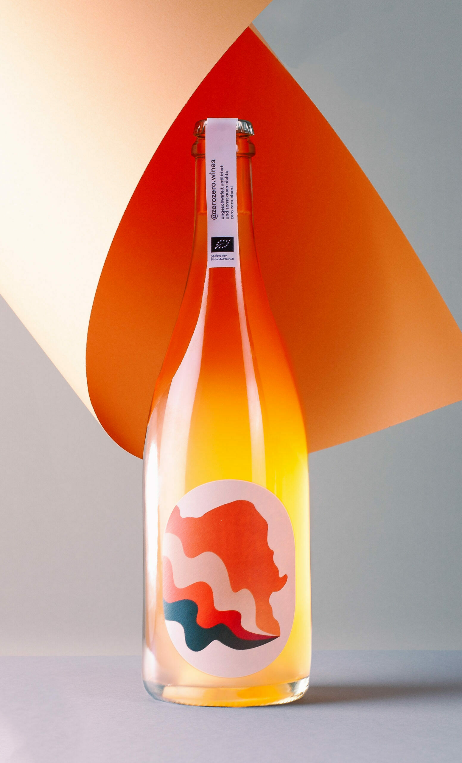 Eine Flasche Pet Nat Orange, die vor einem grau-orangenen Hintergrund steht.