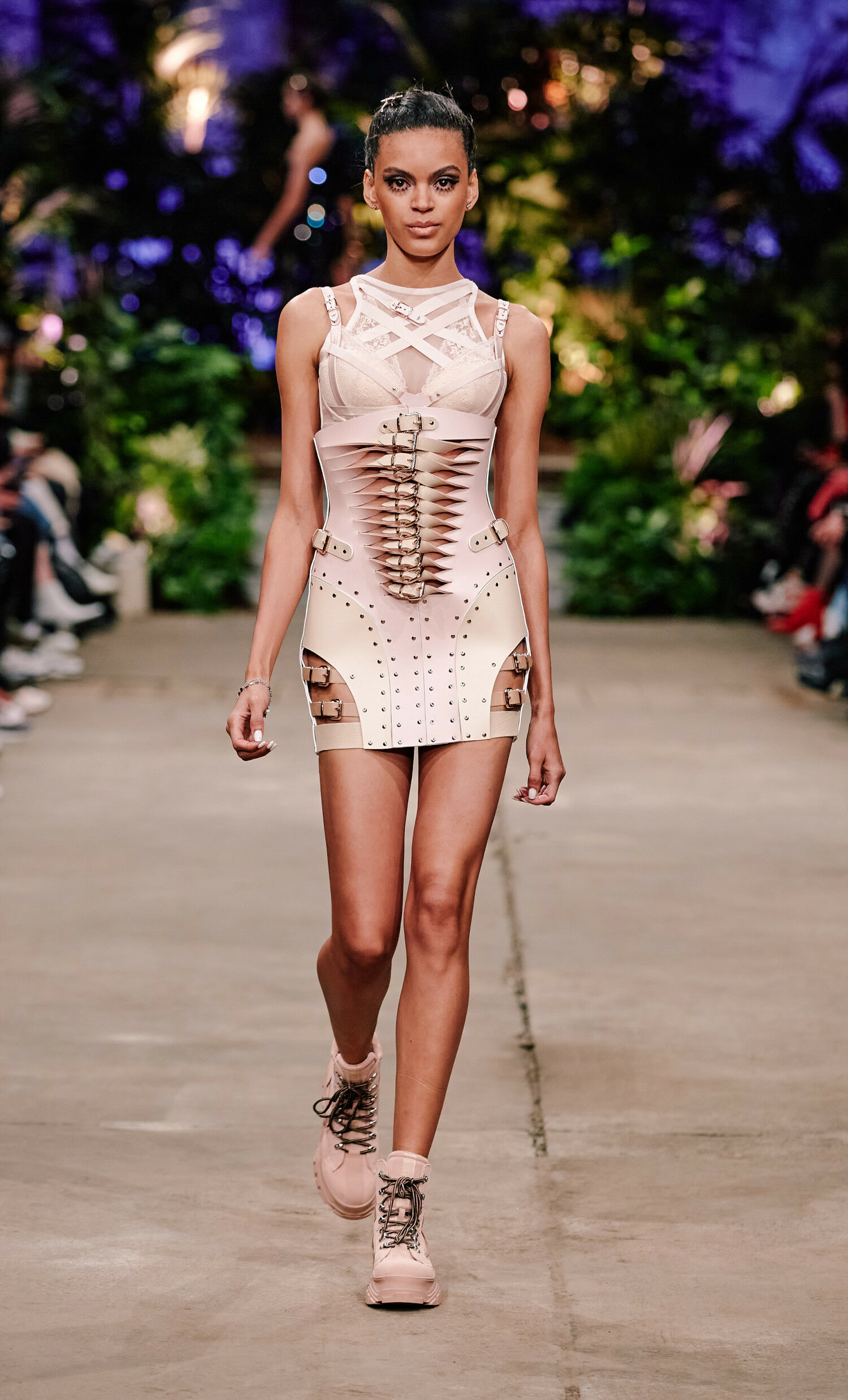 Ein Model in einem enganliegenden Kleid aus festem Material in zartem Rosa läuft über einen Catwalk