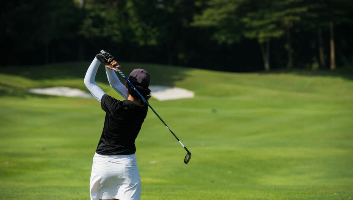 Golferin von hinten holt zum Schlag aus und sielt Richtung Topfbunker vor Bäumen.