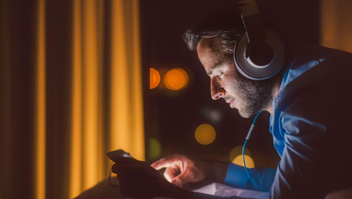 Ein Mann mit Kopfhörern und mobilem Gerät im Liegen, sein Gesicht erleuchtet vom Kunstlicht in der Dunkelheit