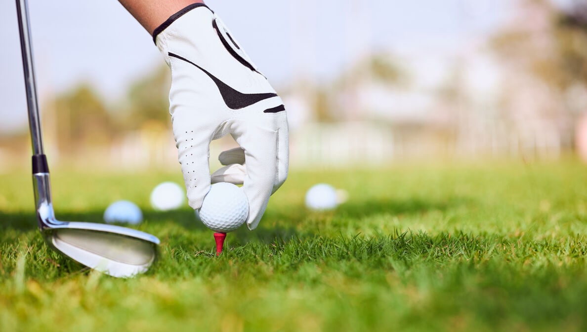 Detailaufnahme von Hand mit Handschuh, die Golfball auf Rasen platziert, daneben Golfschläger