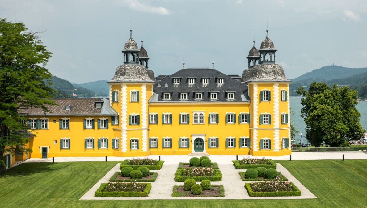 Ein gelbes Schlosshotel vor dem Panorama des Wörthersees