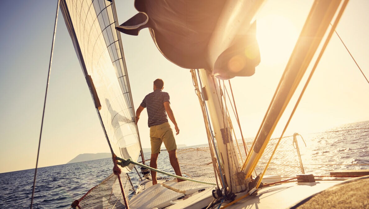 Ein Mann steht am Bug einer Jacht, die bei Sonnenuntergang auf dem Meer in Richtung einer Insel segelt