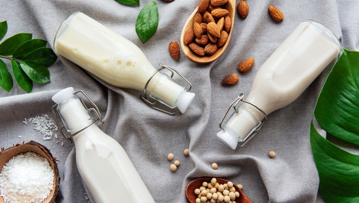 Auf einem grauen Tuch liegen drei Bügelflaschen mit weißlicher Flüssigkeit, Kokosraspel, Kichererbsen und Mandeln