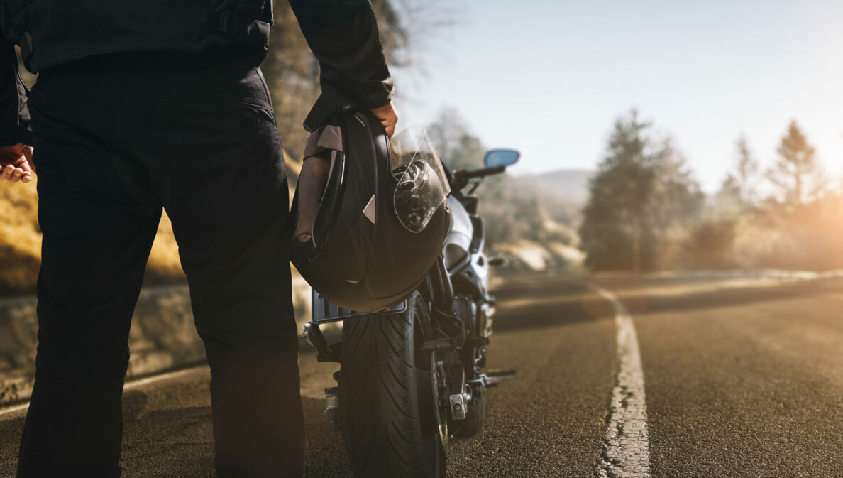 Ein Mann mit Motorradhelm in der Hand steht auf einer einsamen Landstraße vor einem Motorrad.