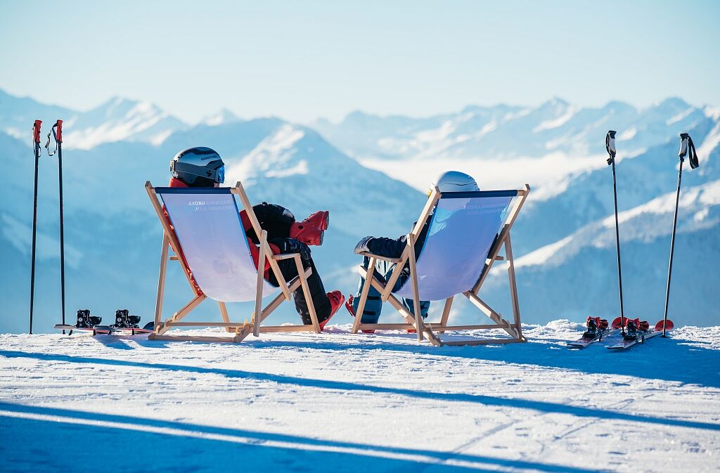 Zwei Menschen von hinten in einem Liegestuhl, sie blicken auf die verschneiten Berge des Skigebiets Wilder Kaiser