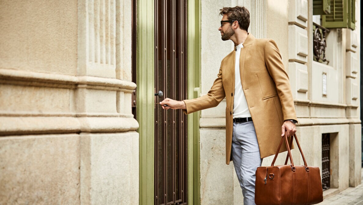 Ein junger, moderner Mann mit Sonnenbrille, beigem Wollmantel und brauner Ledertasche schließt eine Haustür auf