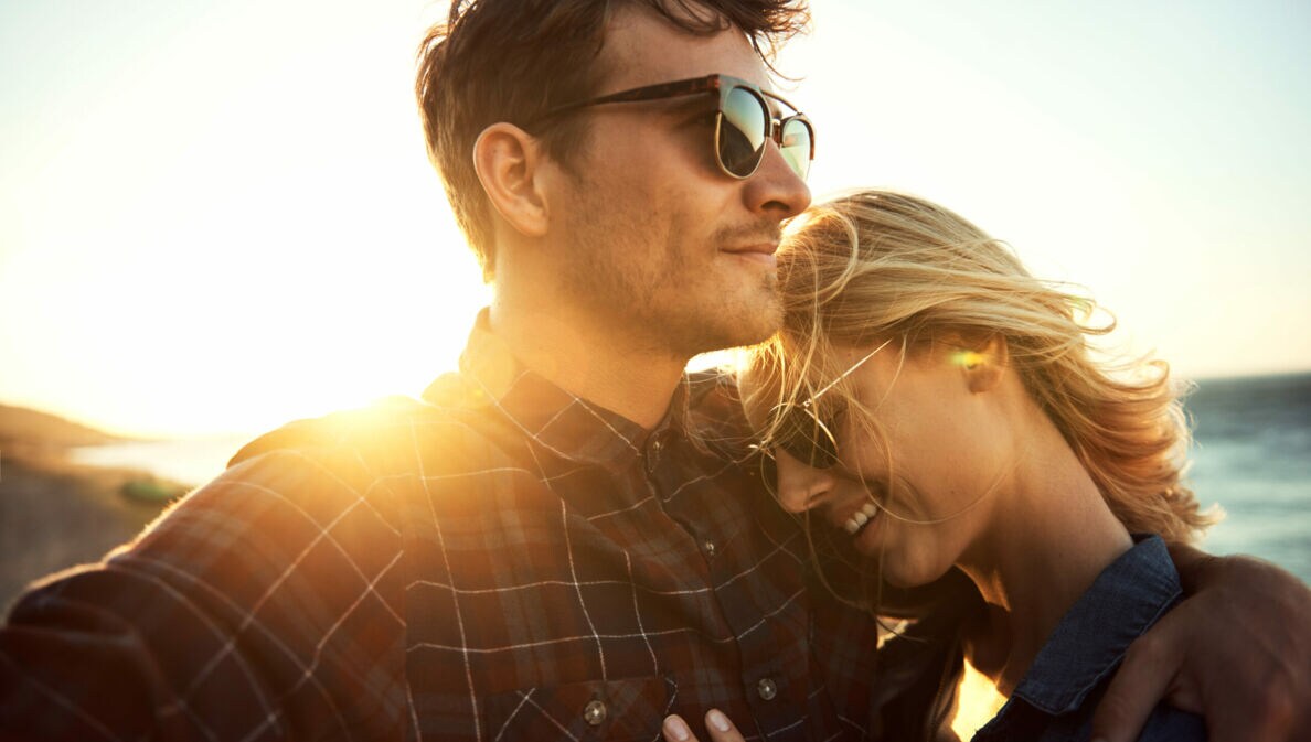 Eine Frau lehnt sich an ihren Partner, sie tragen beide eine Sonnenbrille, im Hintergrund sind das Meer und die untergehende Sonne zu erkennen