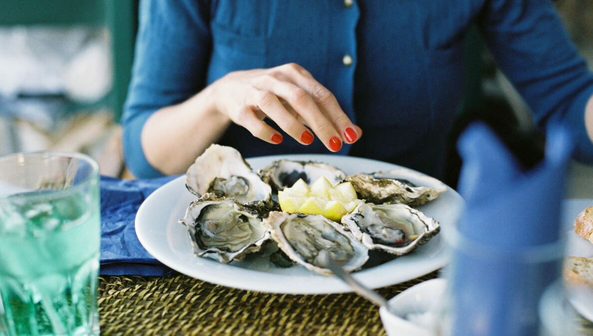 Teller mit Austern auf einem gedeckten Tisch, im Hintergrund Oberkörper und Hand einer Frau