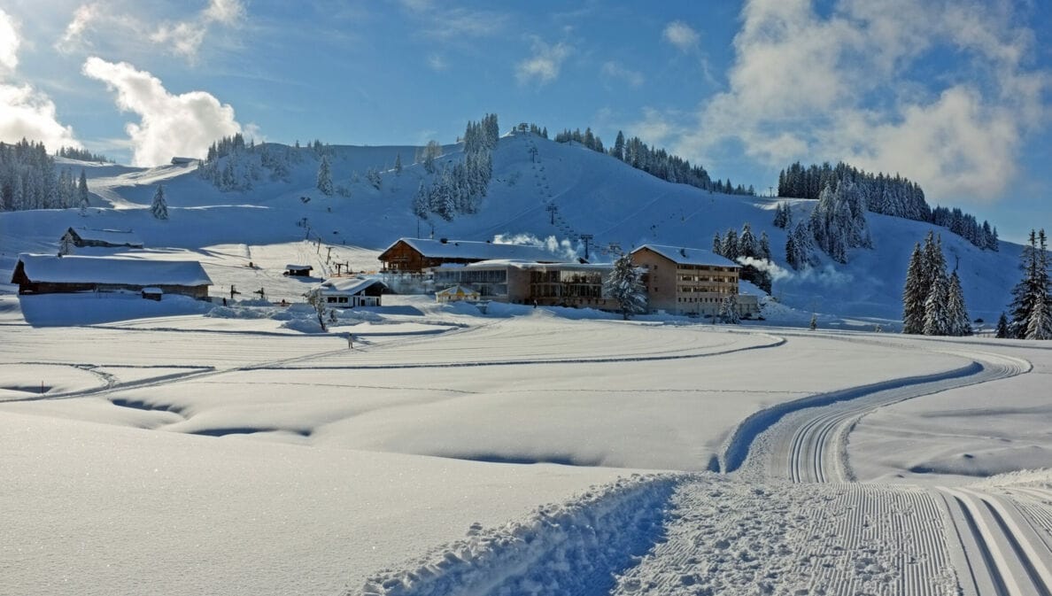 Loipen neben Neuschnee, im Hintergrund Hütten und ein schneebedeckter Berg.