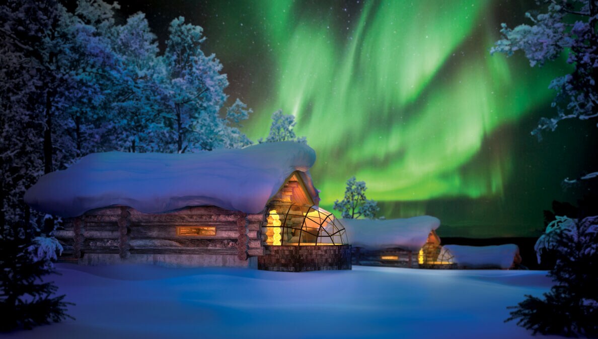 Zwei erleuchtete und schneebedeckte Blockhütten mit Glasanbau in Winterlandschaft mit grünem Polarlicht am nächtlichen Himmel