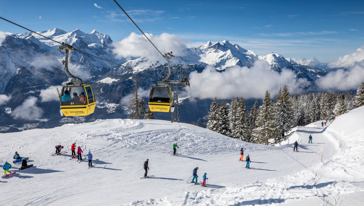 Skipiste mit Wintersportler:innen über die eine Seilbahn läuft, im Hintergrund Bergpanorama