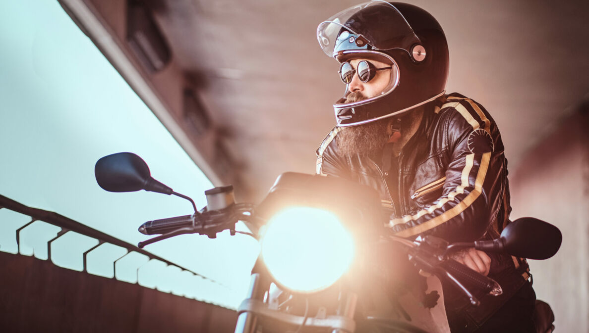 Ein junger, bärtiger Mann in Lederjacke lehnt lässig mit Helm und Sonnenbrille auf einem Motorrad und blickt aus einem offenen Betonbauwerk in die Ferne