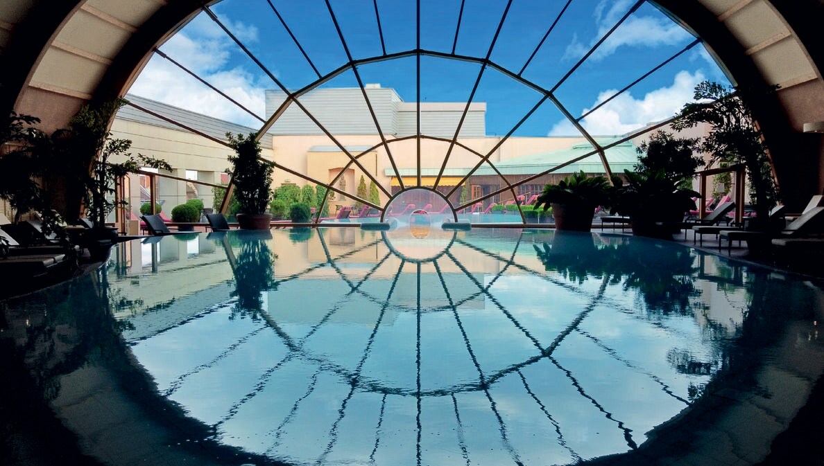Ein großer Innenpool führt durch ein großes, rundes Panoramafenster in den Außenbereich mit Terrasse