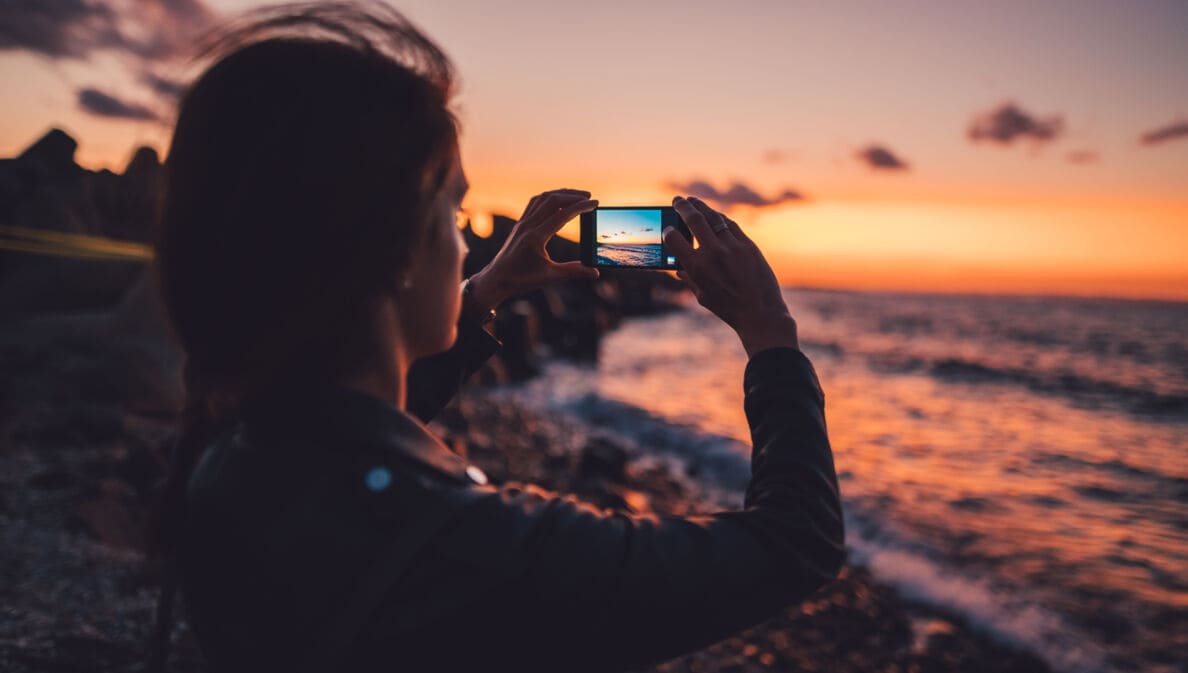Eine junge Frau fotografiert einen Sonnenuntergang am Meer mit dem Handy