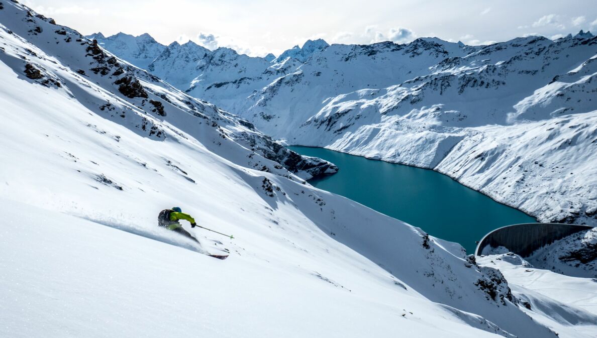 Eine Person auf Ski fährt eine Piste hinunter, im Hintergrund eine Bergkulisse.