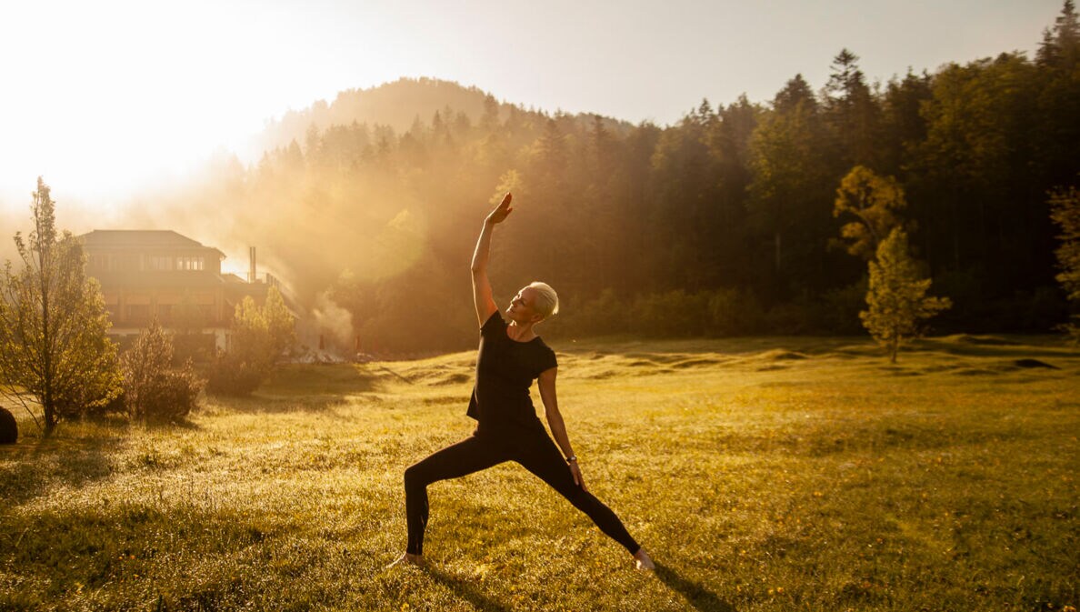 Eine Frau macht Yoga bei Sonnenaufgang und noch leichtem Nebel in einem grünen Garten