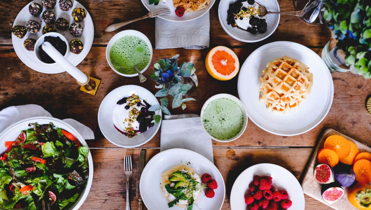 Topshot von unterschiedlichen vegetarischen Speisen auf weißem Geschirr auf Holztisch, umgeben von Besteck, Servietten und Pflanzen