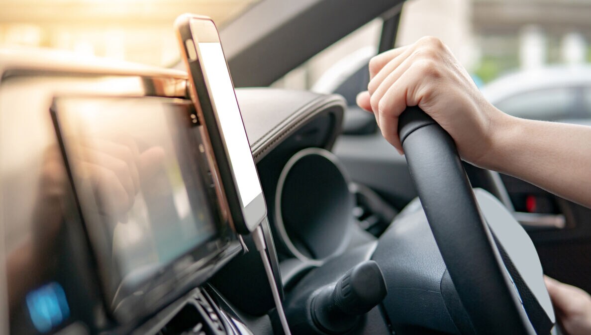Ein Smartphone in einer Halterung am Armaturenbrett eines Autos, daneben zwei Hände an einem Lenkrad
