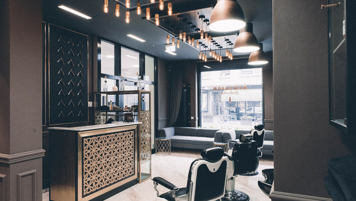 Das Interieur eines modernen Barbershops mit einem Messing verkleideten Tresen, Friseurstühlen aus Leder und einem dunkel gehaltenen cleanen Ambiente