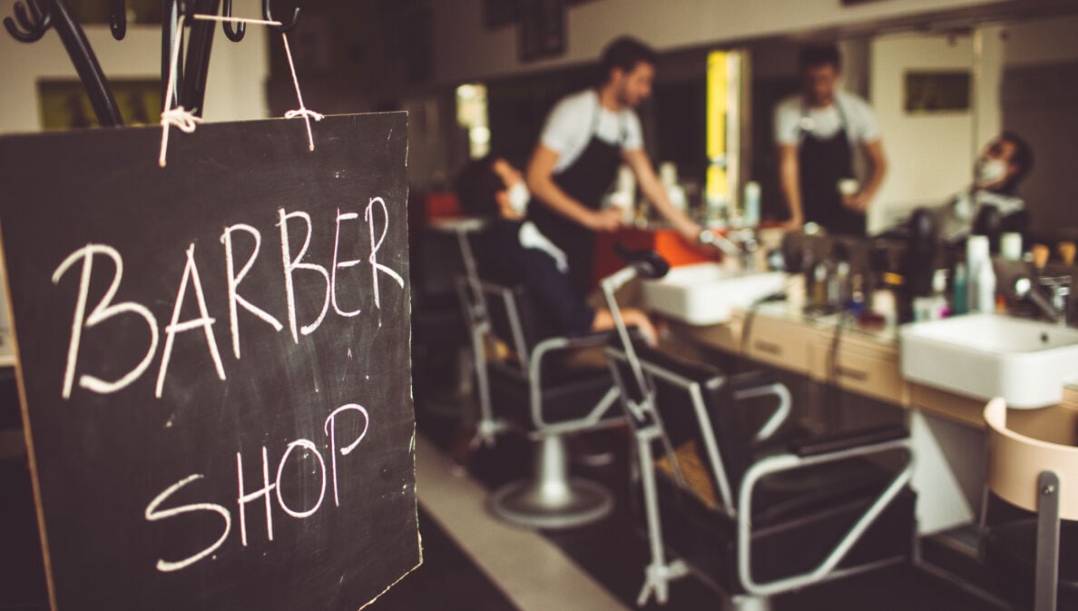 Eine Tafel auf der mit Kreide Barbershop steht, im Hintergrund ist ein Barbier mit seinem Kunden im Laden zu sehen