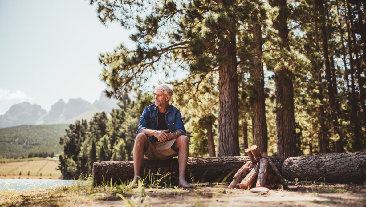 Mann sitzt auf einem Baumstamm, im Hintergrund ein See und Wald