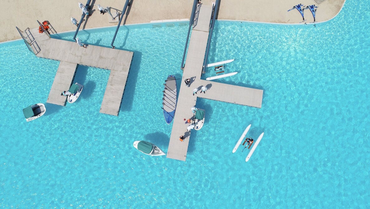 Ein großer Pool mit hellblauem, klaren Wasser sowie Booten, Katamaranen und zwei geometrischen hellgrauen Stegen in Betonoptik