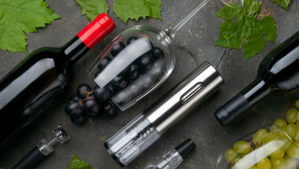 Aufsicht von elektrischem Metall-Korkenzieher, Weinbbelüfter und Vakuumstopfen neben Weinflaschen, Gläsern und Trauben auf grauem Betonhintergrund