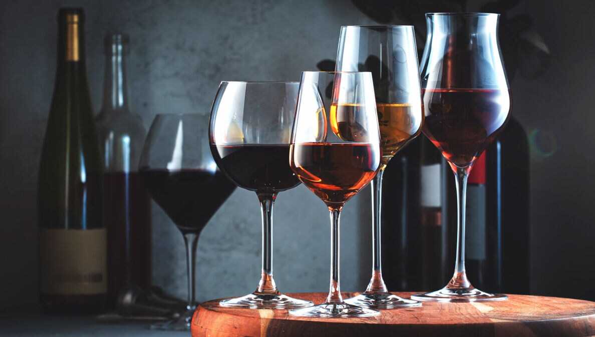 Mehrere gefüllte Weingläser stehen nebeneinander auf einem Brett, dahinter Weinflaschen vor dunklem Hintergrund.