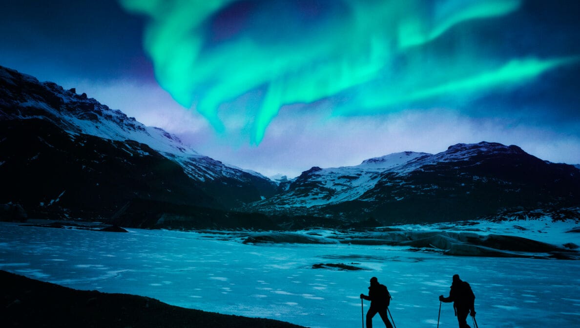 Zwei Wanderer vor einer Bergkulisse mit Polarlichtern.