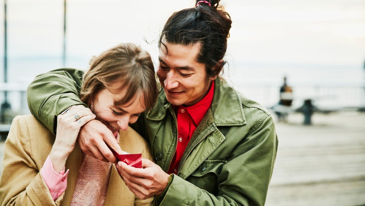 Ein lachendes, sich umarmendes Paar schaut gemeinsam auf eine kleine, rote Geschenkschachtel