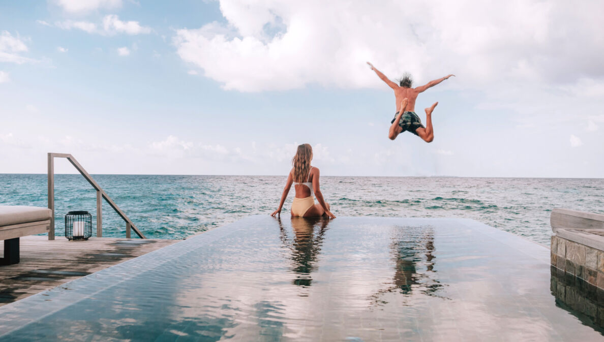 Eine Frau sitzt am Rande eines Pools am Meer, während ein Mann ins Meer springt