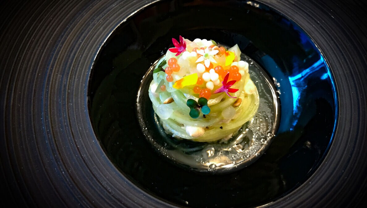 Nahaufnahme eines Gourmet-Gerichtes mit grünen, zusammengerollten Nudeln, dekoriert mit Kaviar und Blüten und angerichtet auf einem schwarzen Teller