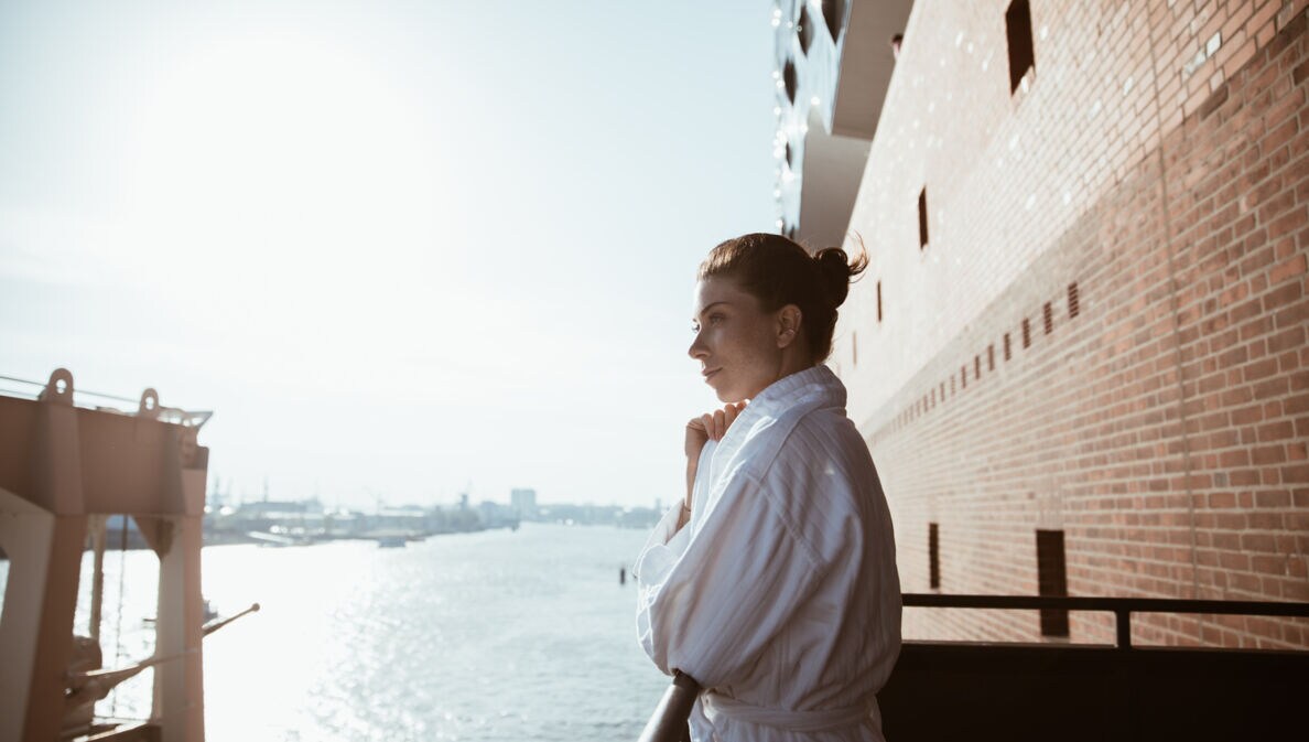 Eine Frau im Bademantel schaut von einem Balkon über die Elbe.