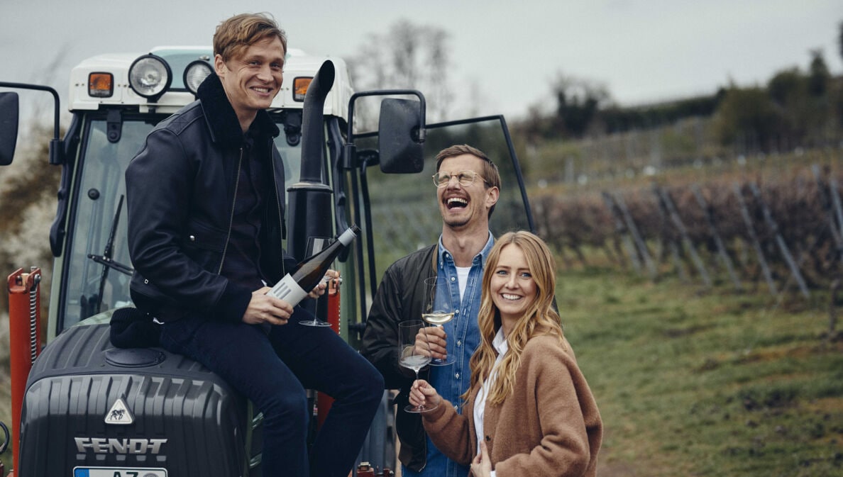 Matthias Schweighöfer sitzt lachend mit einer Weinflasche auf einem Traktor, daneben Joko Winterscheidt und Winzerin Juliane Eller auf einem Weinberg