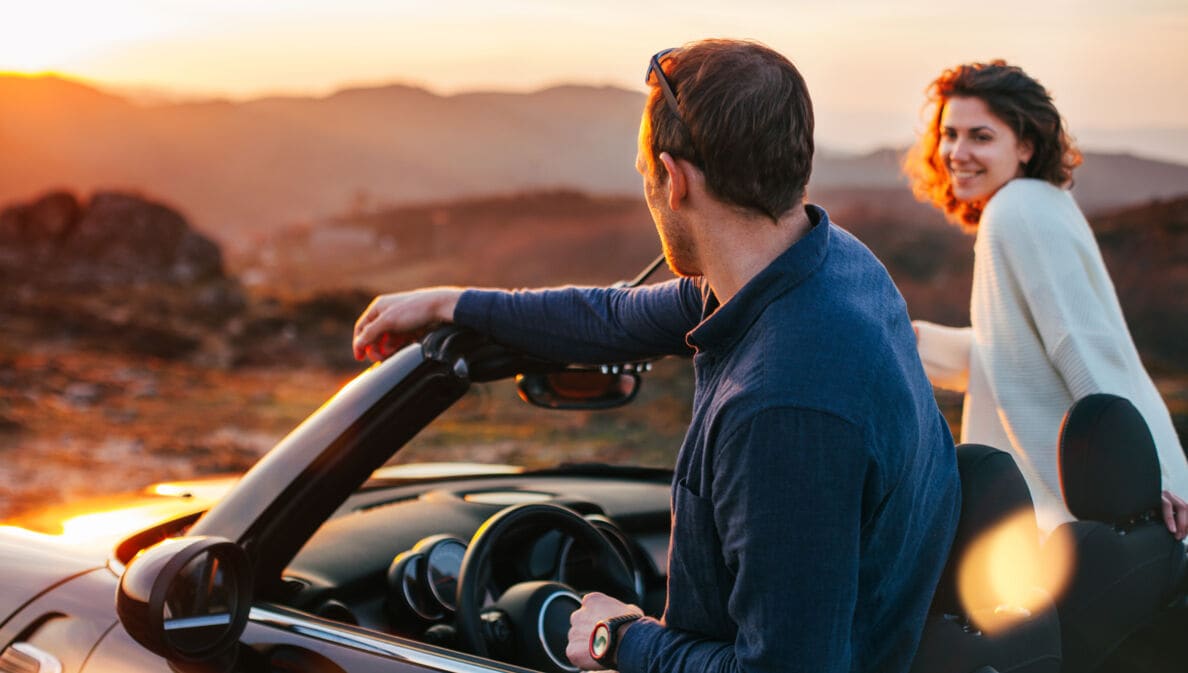Rückansicht eines Paares auf Reise, das aus einem Cabriolet in eine Wüstenlandschaft bei Sonnenuntergang schaut