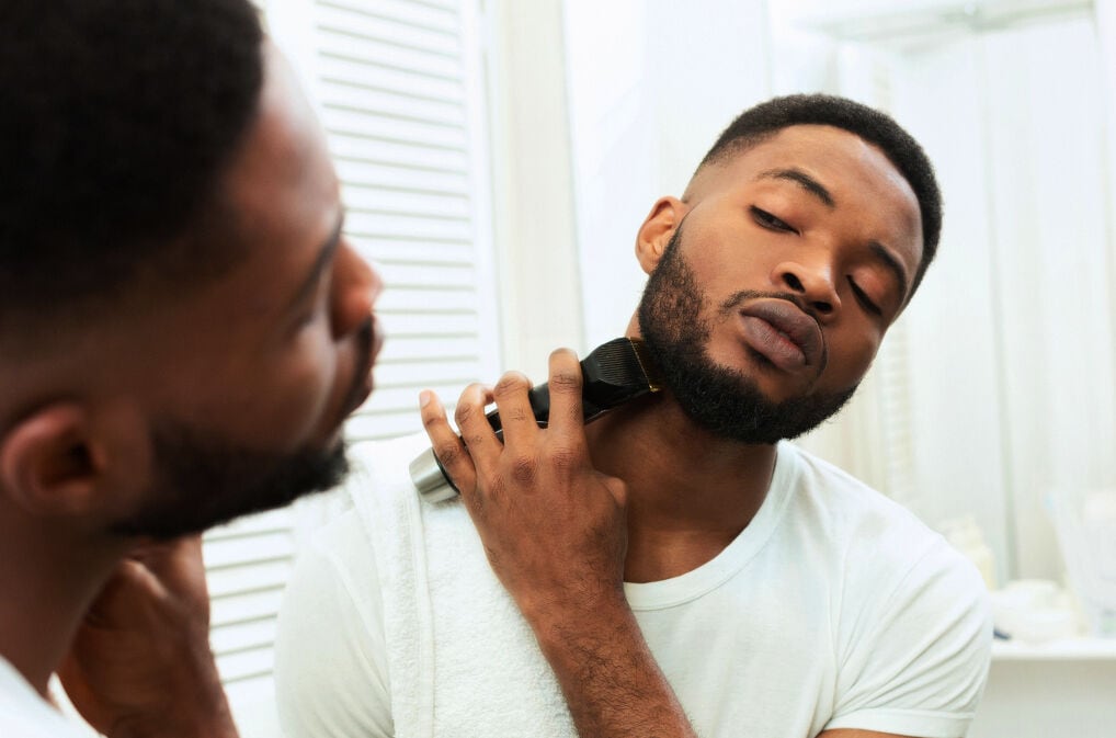 Ein Mann steht vor einem Spiegel und trimmt mit einem Barttrimmer seinen Bart