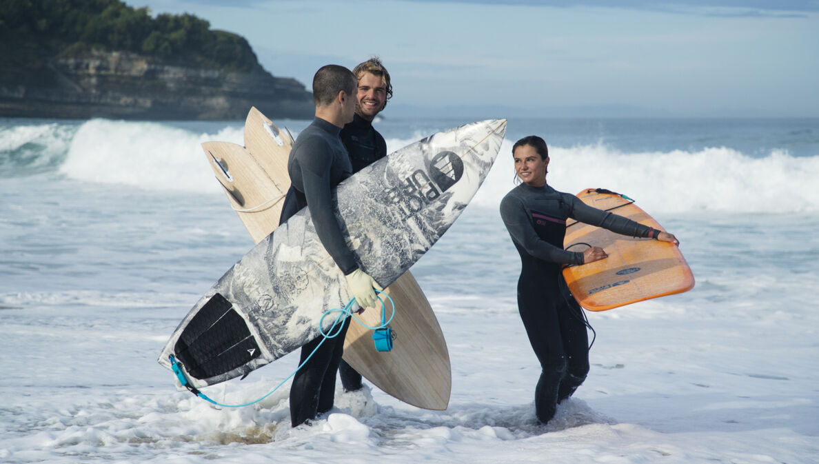 Drei Menschen stehen in Neoprenkleidung und Surfbrettern im Wasser.