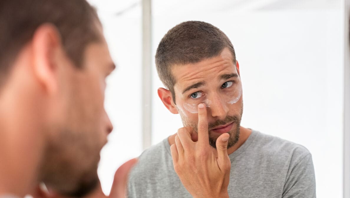 Ein Mann blickt in den Spiegel, während er sich unter den Augen Creme aufträgt