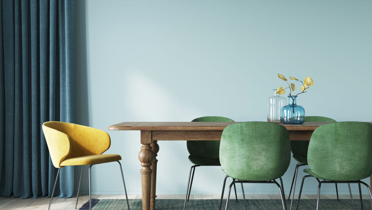 Ein modernes Esszimmer mit Holztisch und Samtstühlen in Sonnengelb und Moosgrün vor einer hellblauen Wand