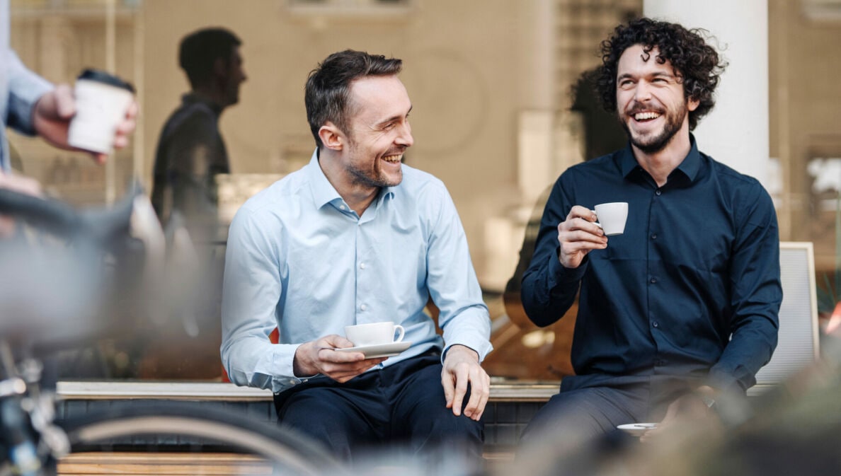 Zwei Männer genießen gut gelaunt einen Kaffee auf einer Bank vor einem Café