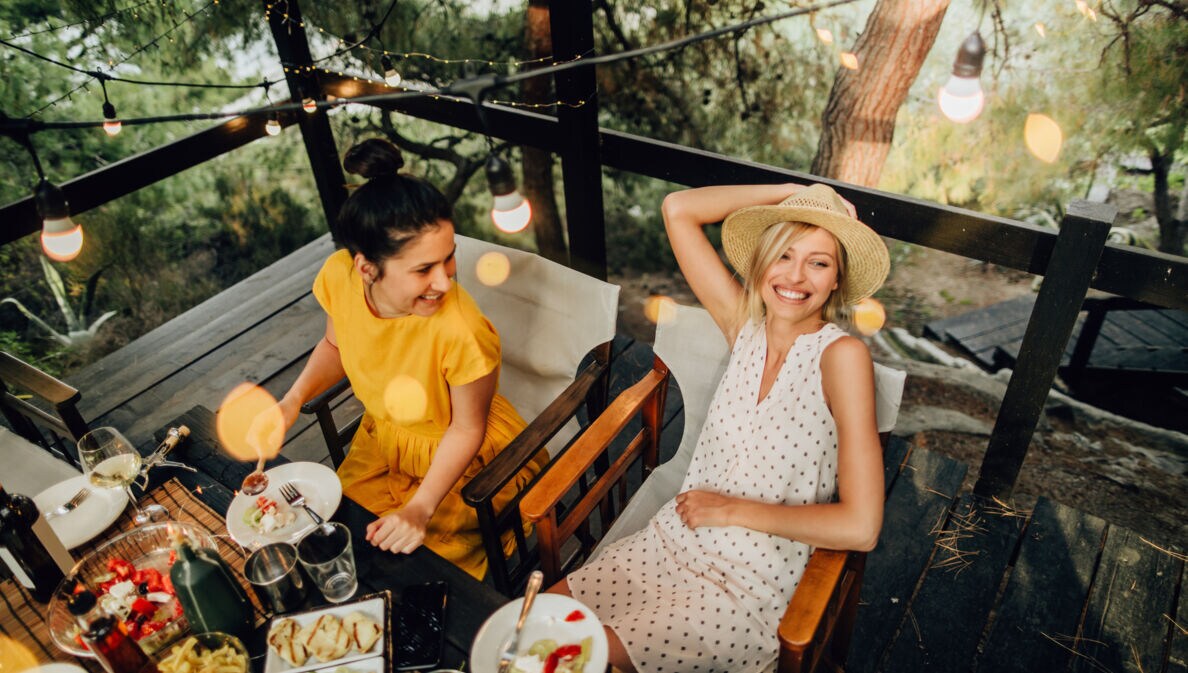 Zwei Frauen sitzen gut gelaunt beim sommerlichen Outdoor-Dinner an einem Tisch auf einer Holzterrasse