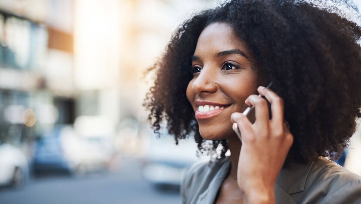 Porträt einer jungen, lächelnden Frau, die mit ihrem Smartphone auf der Straße telefoniert