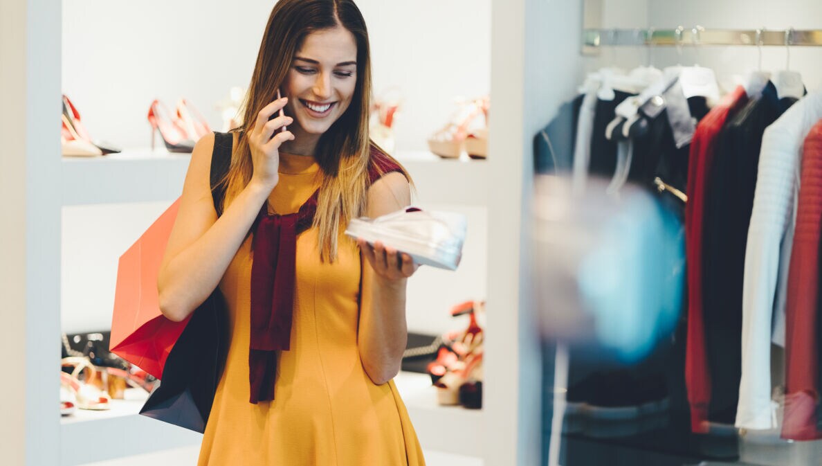 Eine junge Frau betrachtet in einer Modeboutique einen weißen Sneaker in ihrer Hand, während sie telefoniert